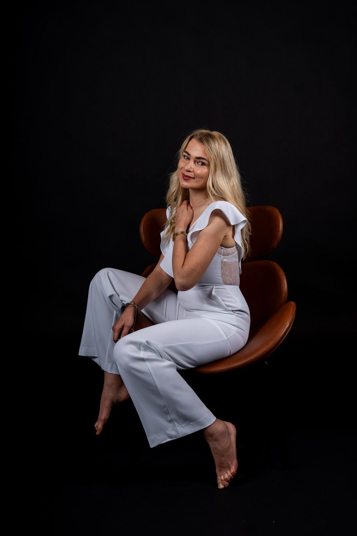 Elegantes Porträt – Weiß auf Schwarz im AuraMirror Studio Frau im weißen Jumpsuit sitzt auf Stuhl vor schwarzem Hintergrund